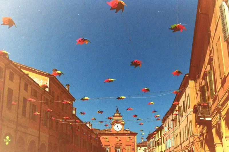 Bright kites crossing a blue sky above a city street lined with old buildings.